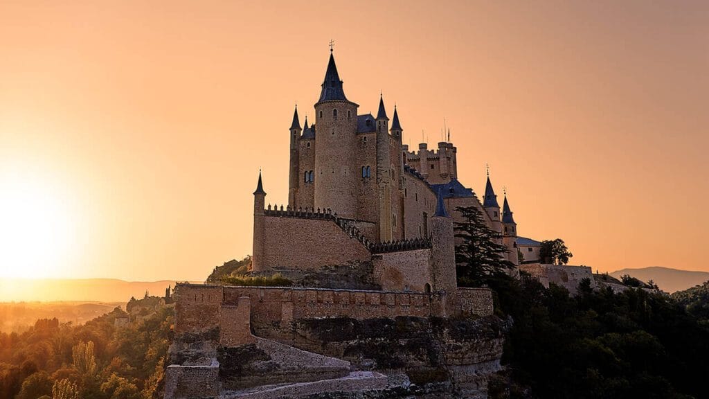 The Alcázar of Segovia rising above the Eresma River with its fairytale turrets and golden stone walls under a clear blue sky.