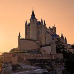 The Alcázar of Segovia rising above the Eresma River with its fairytale turrets and golden stone walls under a clear blue sky.