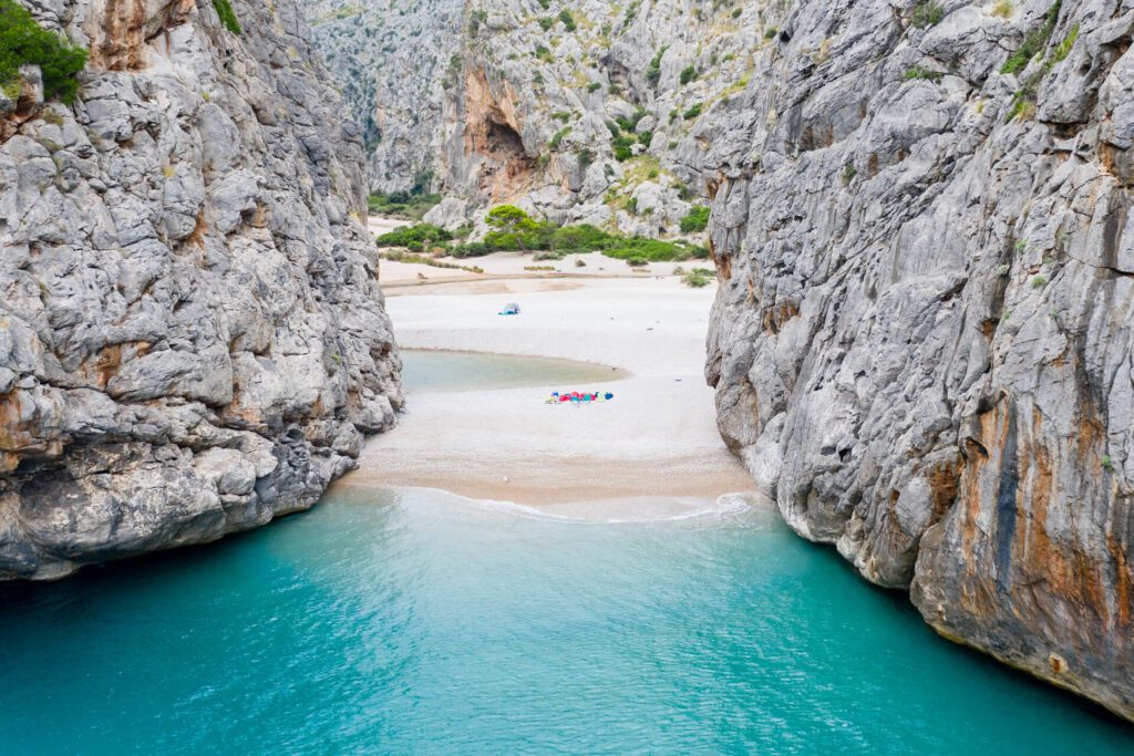 A breathtaking view of Cala Sa Calobra in Mallorca, Spain, a small pebble beach nestled at the end of a dramatic gorge with towering limestone cliffs and deep turquoise Mediterranean waters.