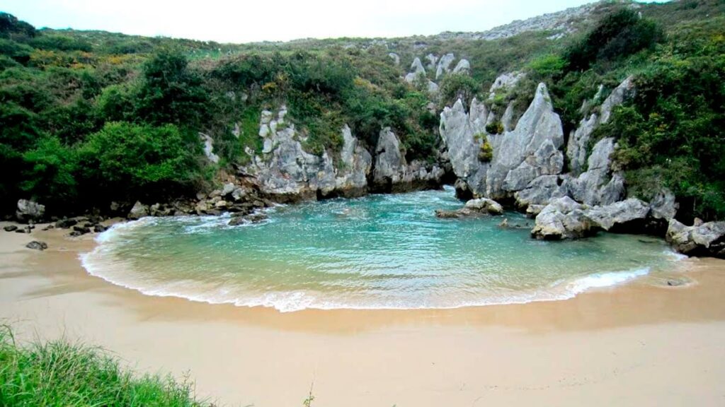 An aerial view of Playa de Gulpiyuri in Asturias, showing the circular inland sinkhole beach surrounded by green meadows and connected to the Atlantic Ocean via underground caves.